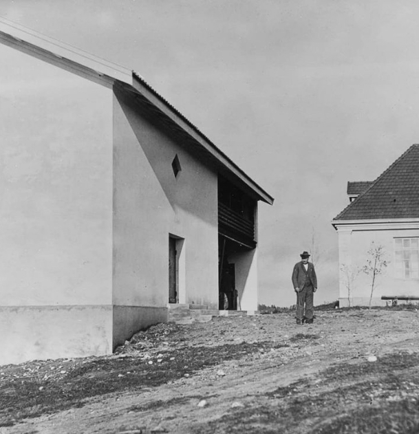 Vanha mustavalkoinen valokuva, jossa mies seisoo kahden vaalean kivirakennuksen välissä. Kevät.